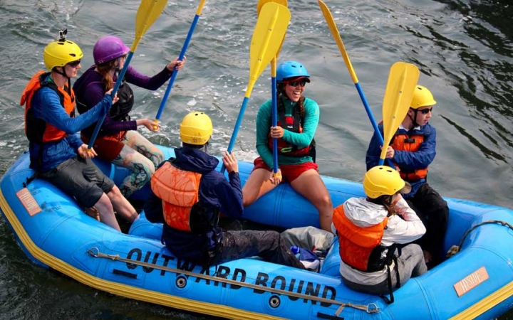 a group of outward bound students wearing helmets and life jackets sit in a raft and raise their paddles into the air
